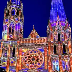 Katedra w Chartres nocą, rozświetlona kolorowymi projekcjami. Architektura gotycka w odcieniach niebieskiego i pomarańczowego.