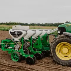 Zielony siewnik do traktorka z białymi zbiornikami na nasiona, gotowy do pracy na polu.