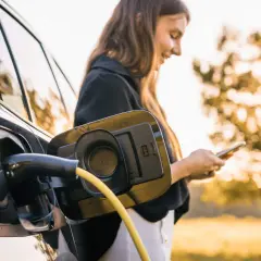 En kvinna laddar sin elbil utomhus, med solen som lyser genom träden. En gul laddningskabel är ansluten till bilen.