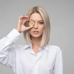 Blondynka w białej koszuli trzyma monetę bitcoin przed okiem, czując zazdrość, że ktoś ma lepiej.