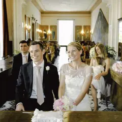 Uśmiechnięty świadek na ślubie, obok panny młodej, w eleganckim czarnym garniturze i jasnoróżowym krawacie.