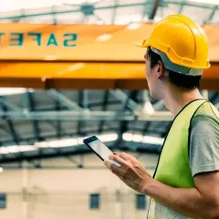 Pracownik w żółtym kasku i kamizelce odblaskowej korzysta z tabletu, dbając o bezpieczeństwo i higienę pracy w hali produkcyjnej.