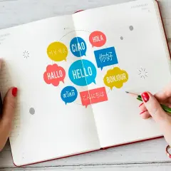 Dłonie nad otwartym notatnikiem, na którym widnieją kolorowe dymki z napisami "HELLO" i powitaniami w różnych językach. To symboliczne przedstawienie **intencji** nauki i komunikacji.