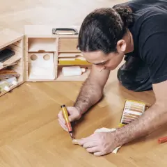 Mann repariert Parkett mit einem Reparaturstift und einer Farbkarte. Werkzeugkasten daneben.