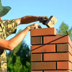 Murarz w zielonej czapce i moro kładzie cegły klinkierowe, używając drewnianego narzędzia.
