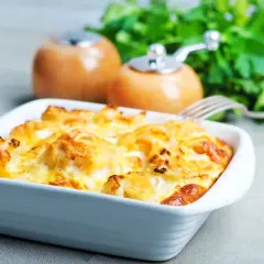Zapiekanka z kalafiora i ziemniaków w białym naczyniu, z widocznym serem i przyprawami w tle.