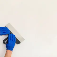Ręce w niebieskich rękawiczkach trzymają szpachelkę, przygotowując się do pracy. Czas, ile schnie gips ceramiczny, zależy od warunków.