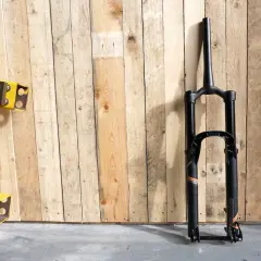 A black MRP Ribbon coil suspension fork stands against a wooden wall. The fork features orange and grey accents and the "ribbon" branding is visible.