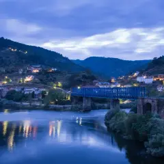 Un puente azul cruza un río tranquilo, reflejando las luces de un pueblo en la ladera. La noche cae sobre este paisaje, creando una atmósfera serena.