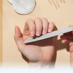 Dłonie pielęgnują paznokcie, używając pilnika. W tle widać narzędzia do manicure, takie jak nożyczki i cążki, tworząc kompletny zestaw do pielęgnacji macierzy paznokcia.