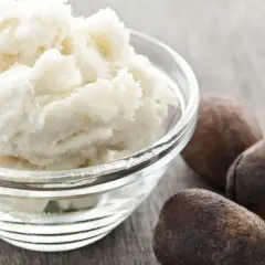 Schüssel mit cremiger Parkii Butter neben drei braunen Nüssen auf Holzhintergrund.