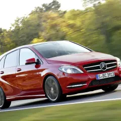 Czerwony Mercedes B Klasa pędzi po drodze, otoczony zielenią drzew.
