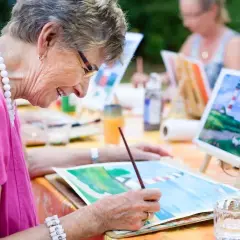 Des personnes âgées participent à des activités manuelles en peignant en plein air. Une dame souriante, vêtue d'un haut rose, est concentrée sur sa toile.