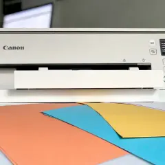 Drukarka Canon TS6351 wyświetla komunikat o błędzie, sugerując, dlaczego z drukarki wychodzą puste kartki. Na biurku kolorowe papiery.