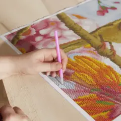 Ręka z różowym aplikatorem umieszcza koraliki do wyklejania na obrazku z rudzikiem i kwiatami.
