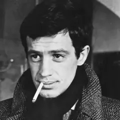 Jean-Paul Belmondo z papierosem w ustach, w płaszczu. Jego spojrzenie przywodzi na myśl niezapomniane **belmondo cytaty**.