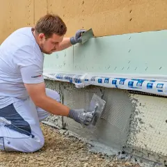 Handwerker kniet und verputzt die Sockelzone einer Hausfassade mit einer Kelle.