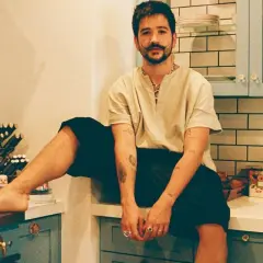 Camilo Alonso Vega, con su característico bigote, posa relajado en una cocina. Sus tatuajes y anillos añaden un toque personal a su estilo.