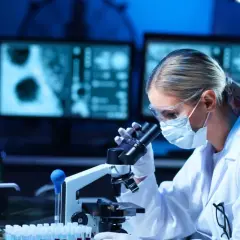 Diagnosta laboratoryjny w okularach i masce analizuje próbki pod mikroskopem. Na ekranach komputera widoczne są wyniki badań.