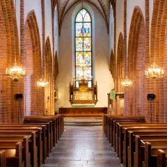 Interiör av Helga Trefaldighets kyrka med bänkar, tegelvalv och ett färgstarkt glasmålat fönster bakom altaret.