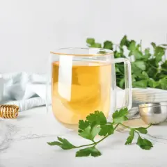 Aromatyczna herbata z pietruszki w szklance, obok świeże gałązki pietruszki, miód i zaparzacz.