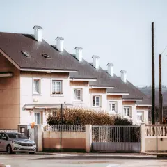 Rządowy dom szeregowy z jasnymi ścianami i ciemnym dachem, z białymi kominami. Przed domem ogrodzenie i samochód.