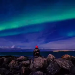 Person sitzt auf Felsen am Meer und blickt auf Nordlichter. Perfektes Wetter auf Island im September für dieses Naturschauspiel.