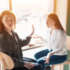 Zwei Frauen mit roten Haaren sitzen sich gegenüber. Eine hält die Hand hoch, als ob sie sagt: "Hör auf zu reden", wenn Kollegen nicht mit einem sprechen.