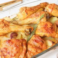 Muslos de pollo al horno con patatas, dorados y jugosos, acompañados de patatas asadas, ajo y romero en una fuente de cristal.