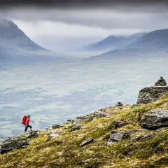 En person med röd ryggsäck vandrar uppför en stenig sluttning med vidsträckt, dimmig fjällvy. Abisko vandring i sitt esse.