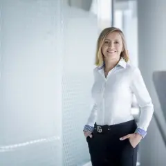 Uśmiechnięta kobieta w białej koszuli i czarnych spodniach stoi w nowoczesnym biurze.