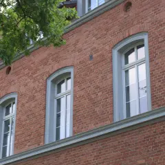 Eleganckie okna drewniane zabytkowe w ceglanej kamienicy, otoczone zielenią drzew.