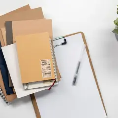 Stos notatników w różnych kolorach, od brązowego po granatowy, leży na białym tle. Obok znajduje się deska z klipsem i długopisem. Całość dopełnia zielona roślina w doniczce. Idealny zestaw do pracy, który inspiruje do tworzenia, nawet gdy masz tylko n...