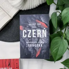 Kolory zła: Czerń. Filmowa okładka książki Małgorzaty Olwii Sobczak, otoczona zielonymi liśćmi i innymi książkami.