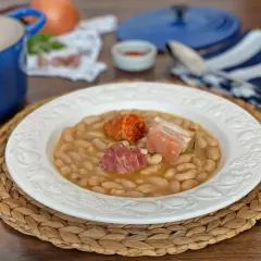 Plato de judías blancas con chorizo y tocino, servido en una vajilla blanca sobre una esterilla de mimbre.