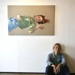 Konstnären Elin Boberg sitter på golvet framför sin målning av en sovande kvinna i grönt mönstrat linne och blå jeans.