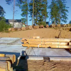 Budowa domu z bali: fundamenty, stosy drewna i pracownik na budowie.