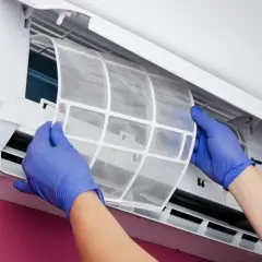 Manos con guantes azules retiran el filtro del aire acondicionado. ¿Para qué sirve? Para atrapar polvo y alérgenos, manteniendo el aire limpio.