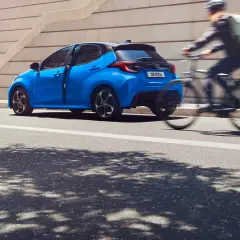 Una Toyota Yaris blu brillante sfreccia in città, mentre un ciclista con zaino passa velocemente. Diversi modelli di Toyota Yaris sono disponibili per ogni stile di vita.