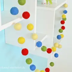 Eine bunte Filzgirlande mit Kugeln in Rot, Blau, Gelb und Grün hängt an weißen Regalen.