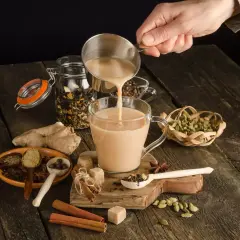 Ręka wlewa mleko do kubka, tworząc aromatyczną herbatę po irlandzku. Wokół przyprawy: cynamon, kardamon, imbir.