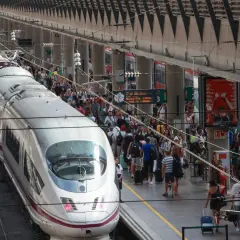 Un tren AVE espera en la estación, listo para llevar pasajeros de Lleida a Sevilla. Gente con maletas se agolpa en el andén.