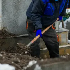 Grabarz w niebiesko-czarnym ubraniu kopie grób łopatą. W tle widać nagrobki i wieńce pogrzebowe.