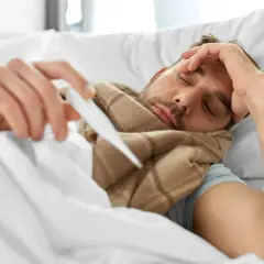 Chory mężczyzna leży w łóżku, trzymając termometr. Zastanawia się, co na gorączkę.