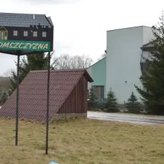 Tablica z napisem "Żeromszczyzna" i stylizowanym domkiem. Obok budynek z napisem "Centrum Edukacyjne Szklany Dom".
