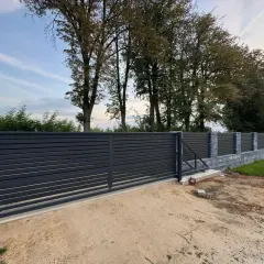 Nowe, nowoczesne ogrodzenie z ciemnoszarymi panelami i kamiennymi słupkami. W tle widać wysokie drzewa w linii ogrodzenia, tworzące naturalną barierę.