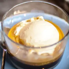 Pyszne affogato: gałka lodów waniliowych w gorącym espresso, podana w szklance na jasnoniebieskim talerzyku.