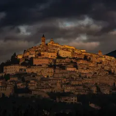 Antico borgo di Trevi, Umbria, illuminato da un sole dorato che filtra tra nuvole scure. Case in pietra si aggrappano alla collina.