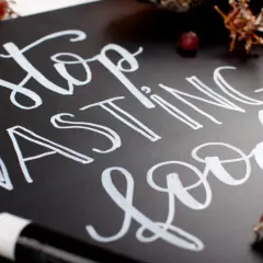 Mensaje "Stop Wasting Food" escrito con diferentes tipos de lettering en una pizarra negra, rodeado de uvas.