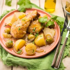 Tajín de pollo con patatas, aceitunas y limón, servido en plato rosa sobre mantel verde.